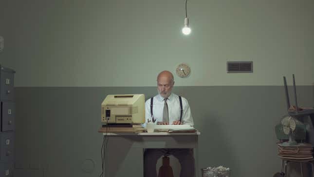 A man sits at a sad desk in a darkened basement with an old computer