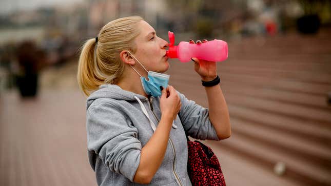 woman pulls down mask to have a drink of water after jogging