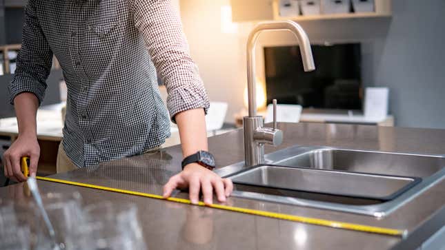 A person wearing a square watch and a navy blue checked shirt uses a yellow tape measure on a dark granite countertop.  