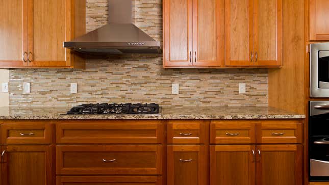 Photo of a kitchen workspace with a gas range, stainless steel hood, brown and gray tiled backsplash, and wooden cabinets. 