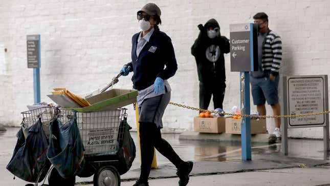 A U.S. Postal Service (USPS) worker wears a mask and gloves while delivering mail, as recipients (R) stand with food they received at a Food Bank distribution for those in need, as the coronavirus pandemic continues on April 9, 2020 in Van Nuys, California.