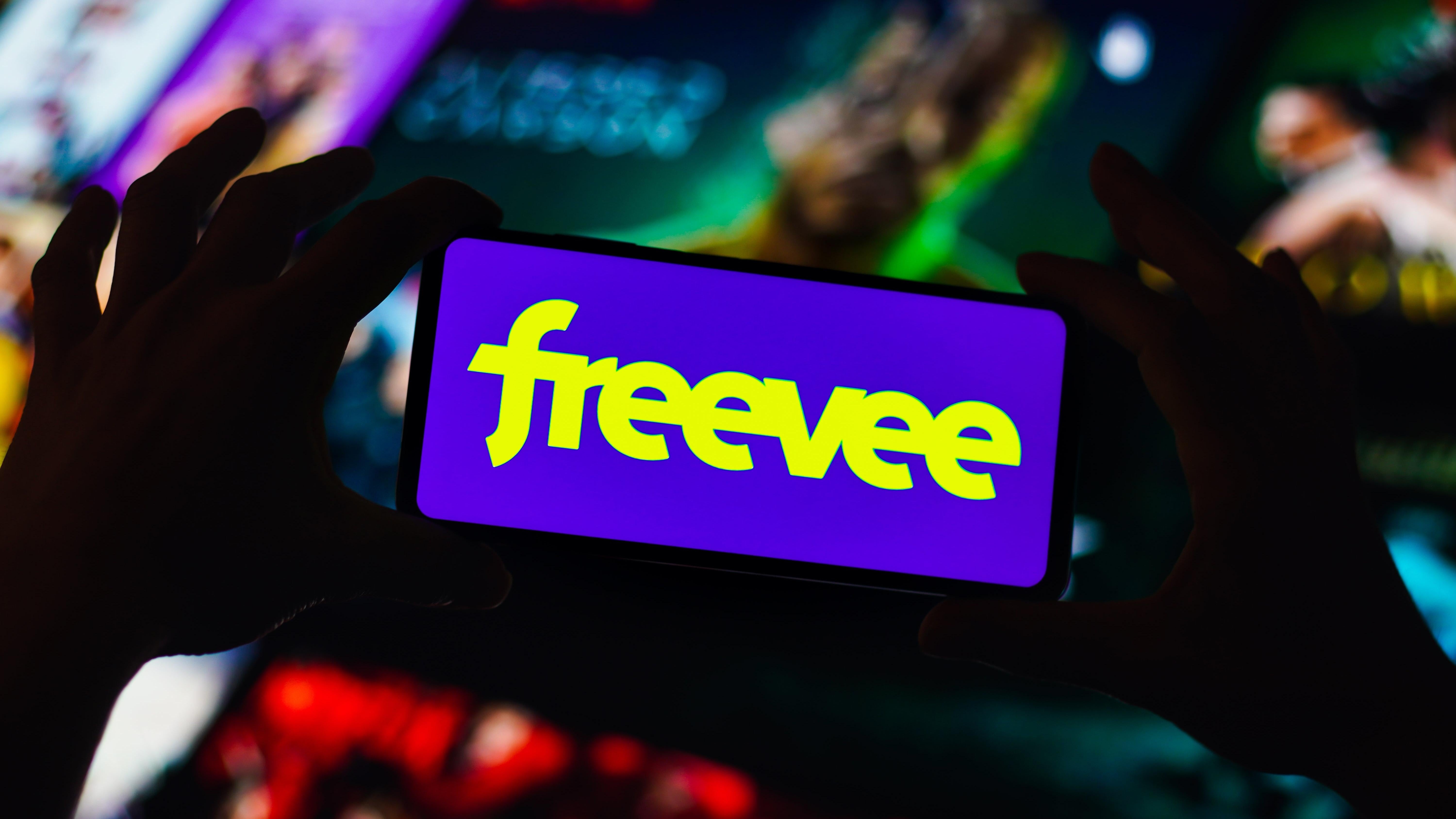 في هذا الرسم التوضيحي للصور ، يتم عرض شعار Freevee على شاشة الهاتف الذكي أمام صف من العروض غير الواضحة.