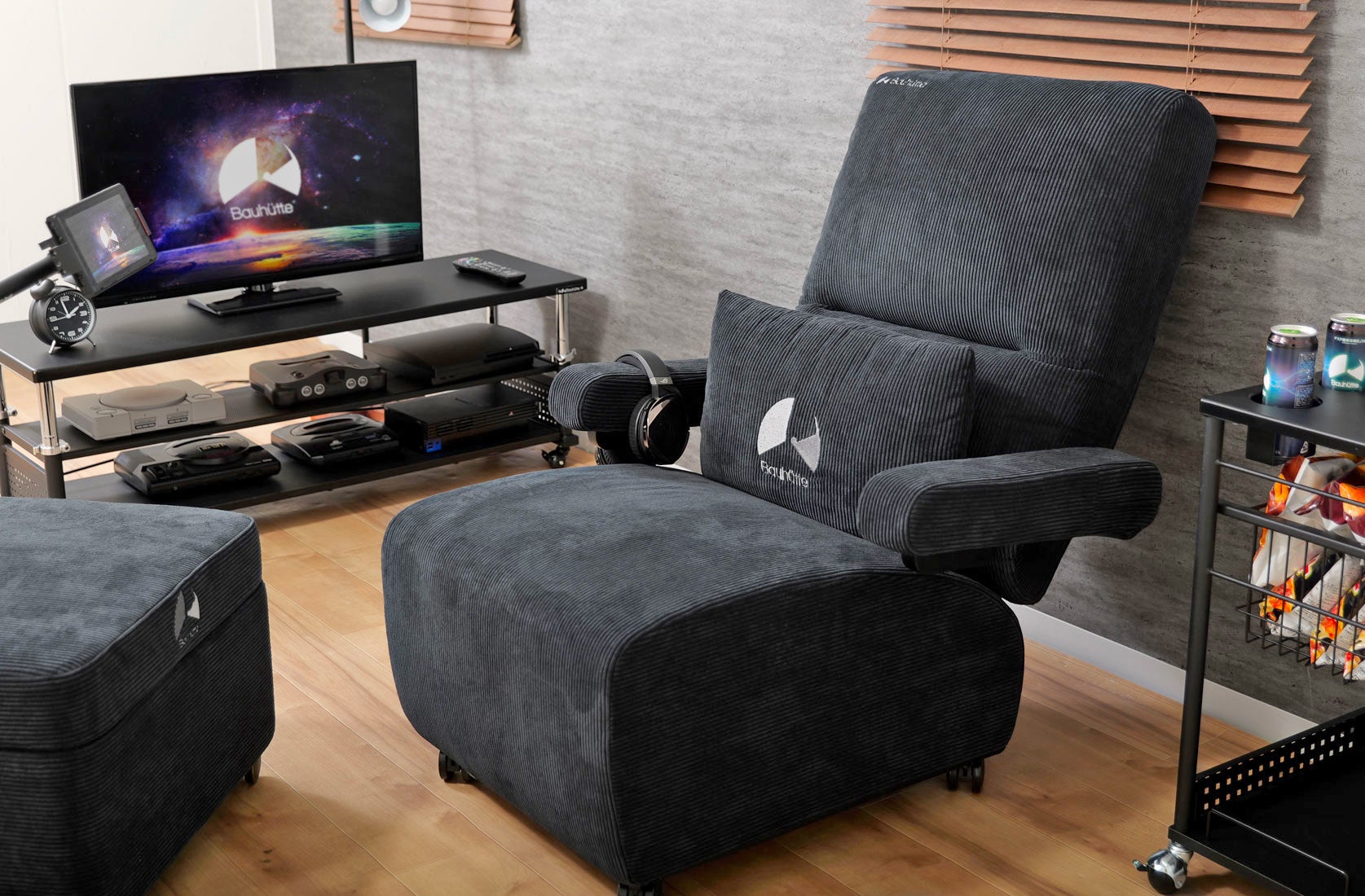 أريكة ديلوكس للألعاب Bauhutte باللون الأسود متوقفة بجوار مركز الترفيه المنزلي الذي يضم العديد من وحدات التحكم.