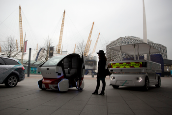 A woman poses for photographers beside a prototype driverless car called a LUTZ (Low-carbon Urban Transport Zone) Pathfinder Pod, center, and a Meridian shuttle, right, during a launch event for the media near the O2 Arena in London