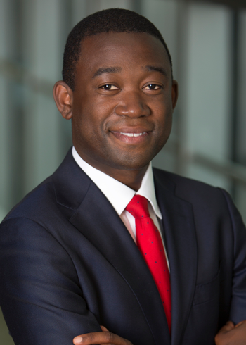 Adewale “Wally” Adeyemo smiles at the camera with his arms across his chest while wearing a dark blue jacket, white dress shirt and red tie.