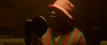 A gif of Wizkid singing into a microphone, wearing sunglasses, an orange hat, and a vest, and dancing in place.