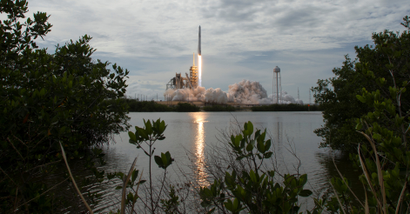 A SpaceX Falcon 9 rocket blasts off from Kennedy Space Center, framed neatly by the sky, sea, and surrounding mangroves.