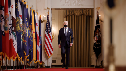 US President Biden walks down the hall to give an address. He is on a red carpet and surrounded by many flags.
