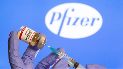 A woman holds a small bottle labeled with a "Coronavirus COVID-19 Vaccine" sticker and a medical syringe in front of displayed Pfizer logo in this illustration taken, October 30, 2020.