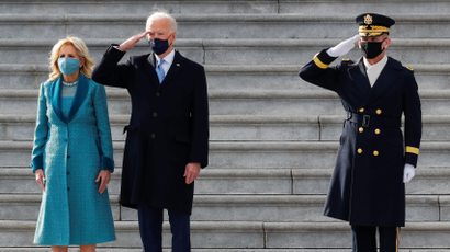 Joe Biden stands on the marble steps of the US Capitol between a saluting marine and First Lady Jill Biden.
