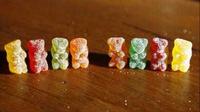 a row of eight gummy bears with sugar coatings. gummies are red, green, orange, and yellow on a wooden table.