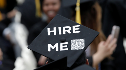 A university student wears a graduation cap with the phrase "Hire Me"