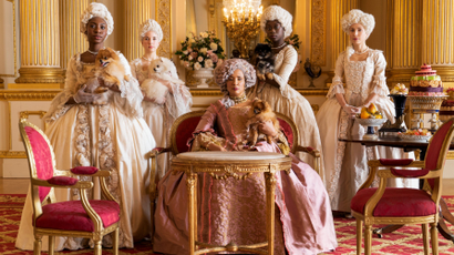 A photograph of the cast of Bridgerton—the most watched show in Netflix history—the cast wear powdered wigs and ornate Regency-era corseted dresses.