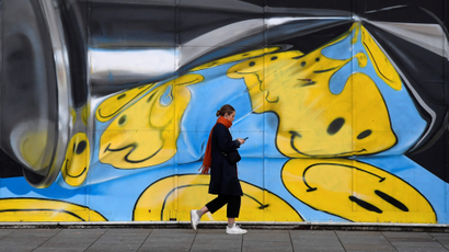 A woman views her phone as she walks past street art of warped emoji smiling faces on a wall in London, Britain,