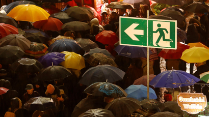 An emergency exit sign rises above the umbrellas of people crowding an outdoor market.