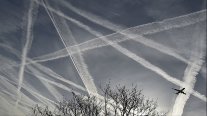 A plane flying through the sky full of chemical trails from previous flights, with an image of a tree in the foreground.