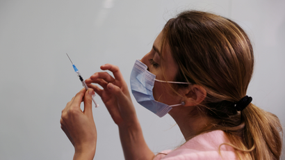 A woman with a mask on holds a syringe of a Covid-19 vaccine. She's wearing pink scrubs.