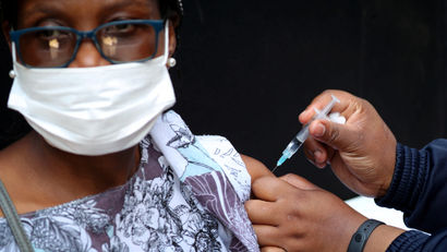 a woman wearing a purple flowered blouse and a white face mask receives a dose of a vaccine in her arm