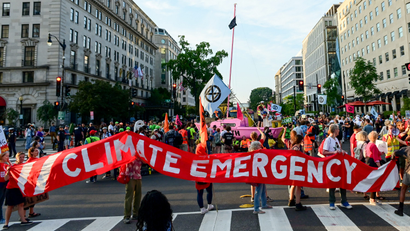 A group of protesters walk down a city street carrying a red banner that reads CLIMATE EMERGENCY