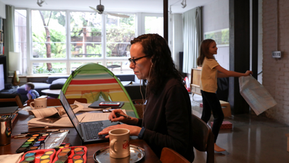 A woman wearing glasses juggles work and childcare responsibilities. She sits at a table covered in paint pallets, papers, two mugs, and other items, trying to work on a laptop in a large multifunction room. A young girl plays in the background on the right and a young boy can be seen on the left in front of a couch. A colorful striped play tent can be seen in the background in front of a large window.