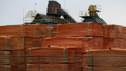 stacks of wood planks are piled up in a lumber yard with processing equipment in the background
