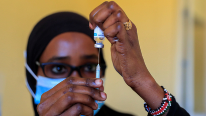 A woman in spectacles, a mask and a black head covering holds a syringe with its needle inside a vial.