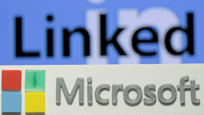 A 3D printed logo of Microsoft is seen in front of a displayed LinkedIn logo over a blue background.