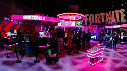 A neon pink sign says "Free Trials" in all-caps in white letters while a larger sign saying Fortnite is in the background at the Epic Games booth at the E3 convention. A shopping cart outfitted with neon pink lights is also in the lower right corner.