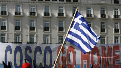 Protesters raise a Greek flag in front of a banner that reads "Cosco Go Home"