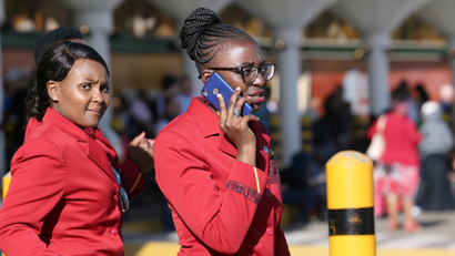 A flight attendant in red attire and glasses talks on her blue mobile phone while another one walks behind her.