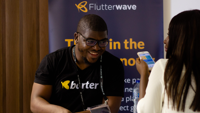 A man laughs in front of a banner for the Flutterwave company. In front of him is a woman holding a phone.