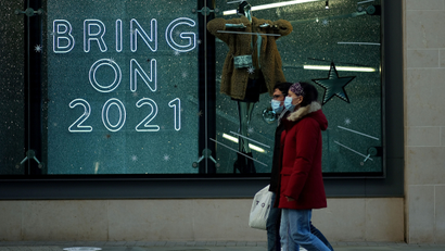 Two people, wearing blue disposable masks, walk past a store with a neon window sign that says "bring on 2021" in large capital letters.
