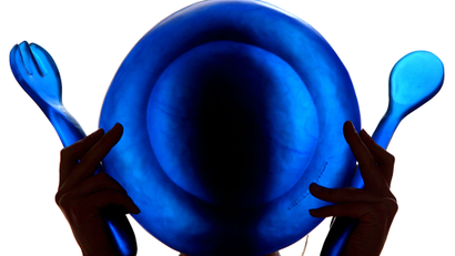A person holds up a translucent blue bowl, fork, and spoon in front of her face.