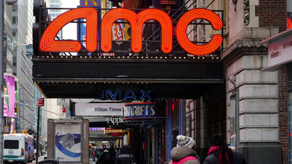 A neon AMC theater sign is pictured while someone walks past wearing a bright red backpack.