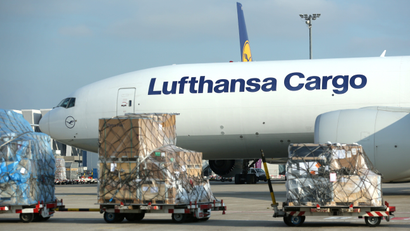 Workers wheel carts laden with boxes down an airport tarmac past an idling Lufthansa cargo plane.