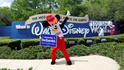 A person wearing a mouse costume dances while holding a Governor Ron DeSantis poster