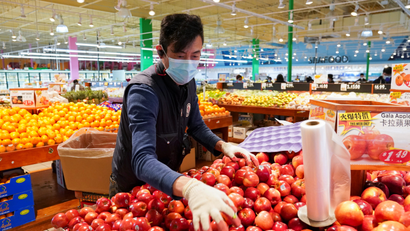Wearing a mask and gloves, a worker re-stocks apples in an Asian grocery store