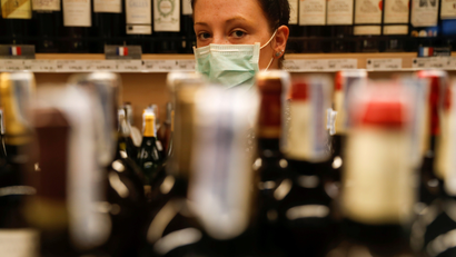 A woman looks at the camera through wine bottles lined up at a grocery store.