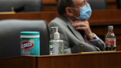 Clorox wipes are featured prominently on a desk with Purell hand sanitizer. A man is wearing a mask in the background.
