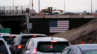 Rows of cars on a highway pass under an overpass with a construction vehicle and an American flag hanging from it