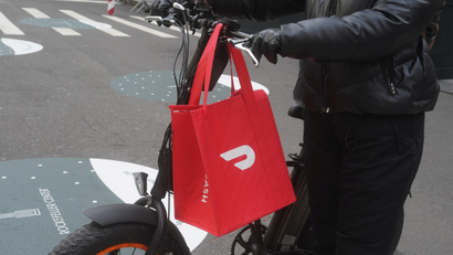 DoorDash delivery worker holding DoorDash-branded bag while on bike outside.