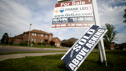 a white and red FOR SALE sign is planted in a patch of grass in front of a brown brick home. Leaning against it is a blue sign that reads SOLD ABOVE ASKING