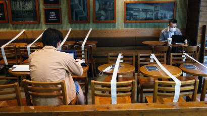 Two people sitting in a cafe. There is white tape separating their tables, sectioning off areas where customers can sit. Each of the people are wearing masks at their respective tables.