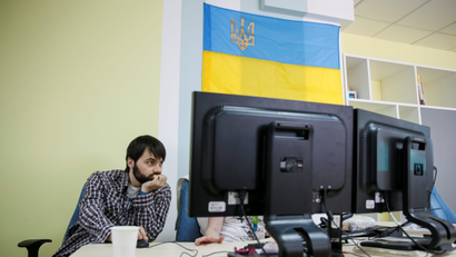 a dark-haired man sits at a desk looking at a computer screen. Above him hangs a national flag of Ukraine that is blue on top and yellow on bottom