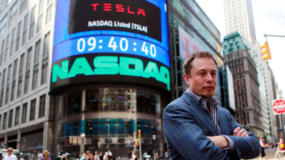 Elon Musk stands with his arms crossed in front of a New York City building with a sign announcing Tesla's listing on the Nasdaq.