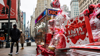 On a grey sidewalk, items sit for sale for Valentine's Day, including a red balloon, a teddy bear, and a white gift bag that reads "Happy Valentine's Day"