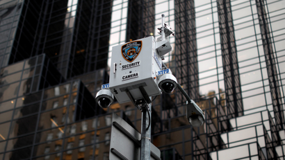 A security camera with New York Police Department logo is seen. A skyscraper is in the background.