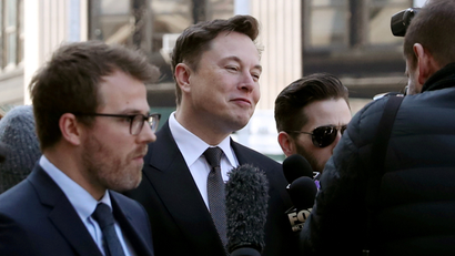 Tesla CEO Elon Musk arrives at Manhattan federal court for a hearing on his fraud settlement, dressed in a black suit and standing in front of a microphone.
