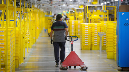 An Amazon worker pulls a cart in a warehouse, passing yellow bins