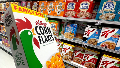 Kellogg's Corn Flakes cereal box in front of cereal aisle of supermarket.